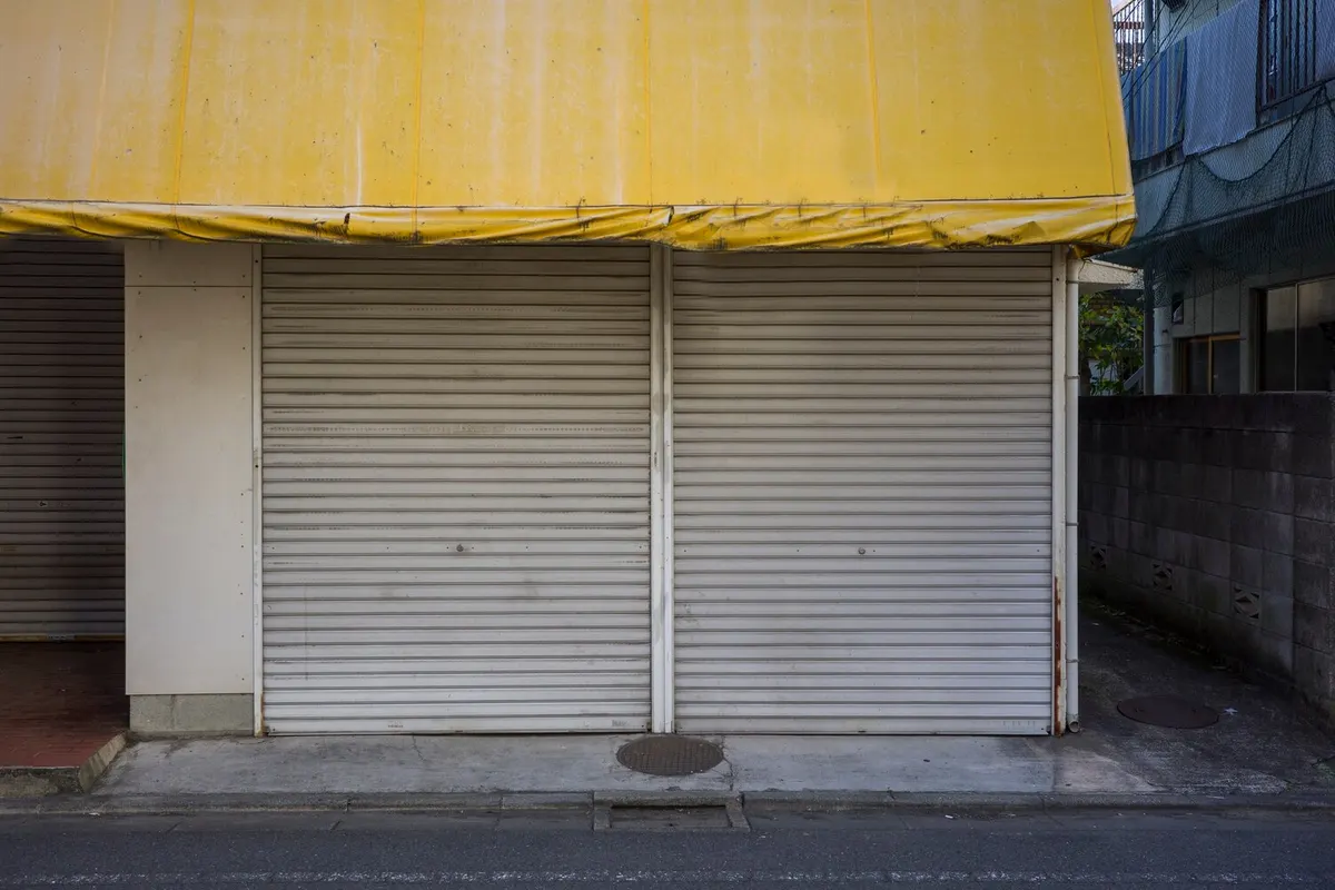 Deux rideaux métalliques gris baissés sous un store jaune délavé, devant une boutique fermée