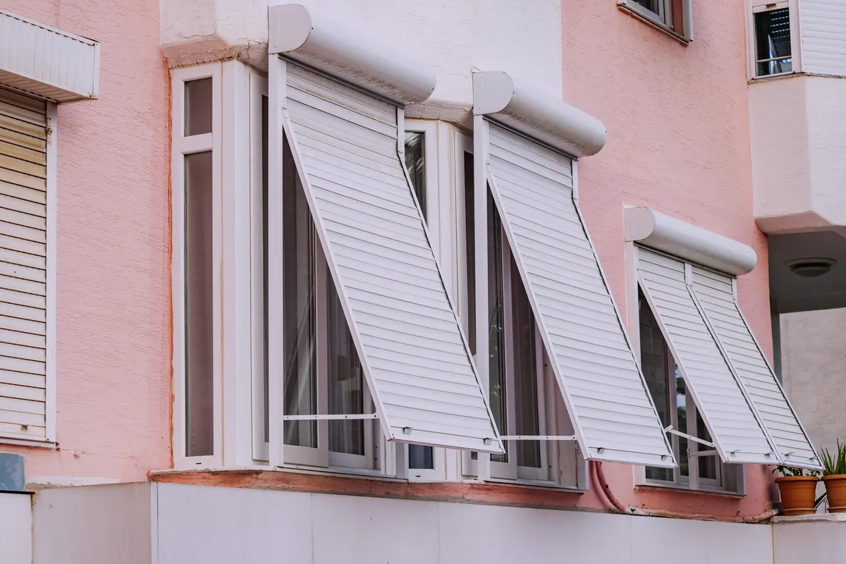 Volets roulants blancs à projection, tabliers inclinés devant les fenêtres d'un immeuble à façade rose